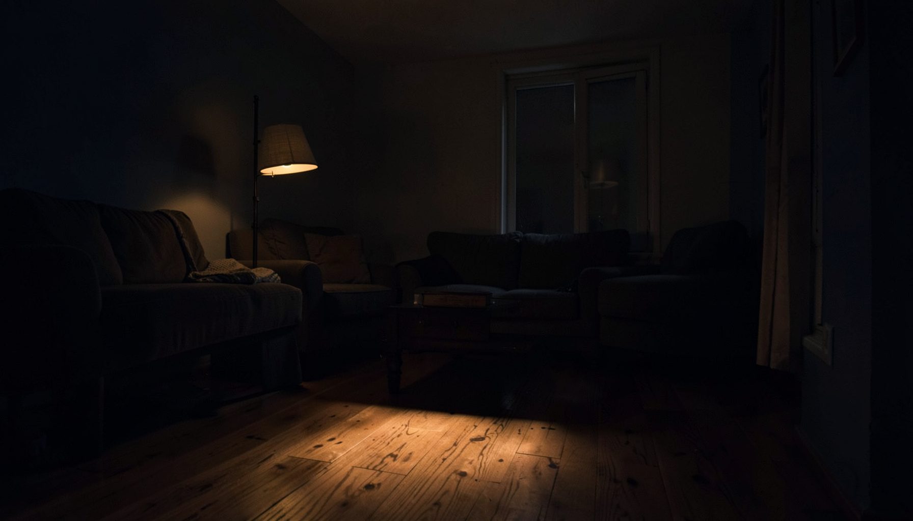 A dim, moody interior of a modest living room with warm amber light spilling across a wooden floor, deep navy shadows in the corners